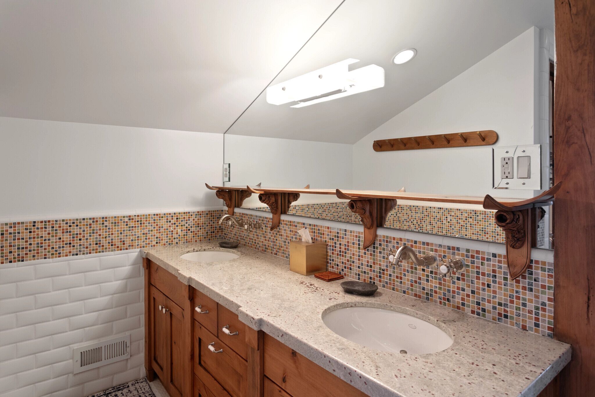Une salle de bain avec deux lavabos, des meubles en bois, un pare-feu en carrelage et un miroir surmonté d’une étagère en bois.