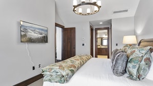 Un dormitorio con una cama, un banco, una araña, un cuadro y una puerta que da a un baño.