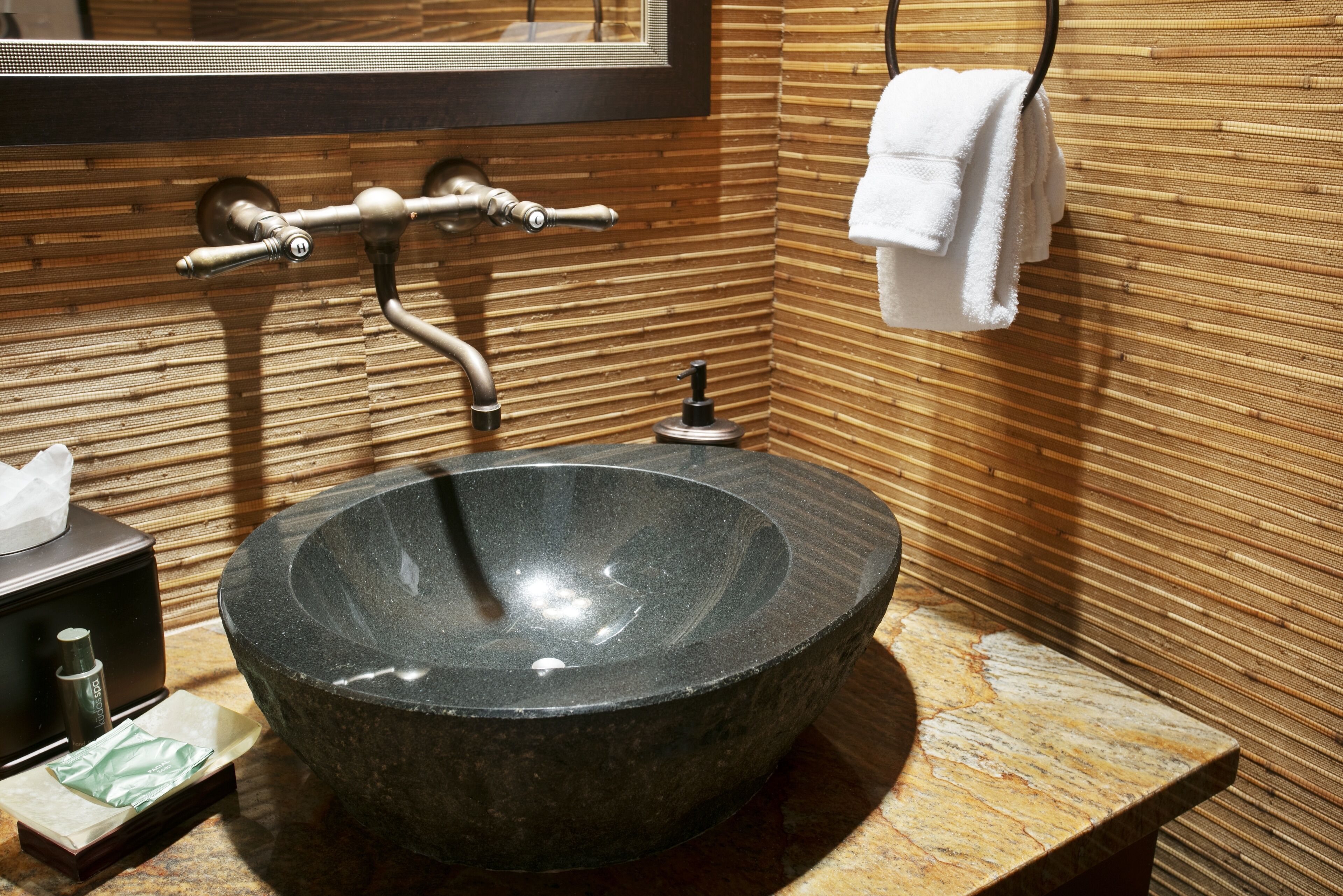 A modern bathroom with a large, round, dark stone sink, a wooden wall with horizontal lines, a towel rack with a white towel, and a mirror above the sink.