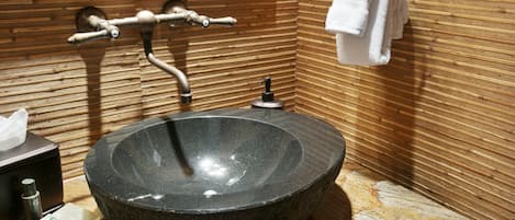 A modern bathroom with a large, round, dark stone sink, a wooden wall with horizontal lines, a towel rack with a white towel, and a mirror above the sink.