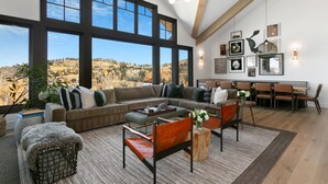 A spacious living room with a large window, a sofa, chairs, a coffee table, and a dining area with chairs and a table. - Tower Villas - Five Bedroom Villa w/ Hot Tub #7700 by Deer Valley Resort (Park City)