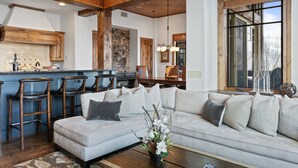 A spacious living area with a large sofa, a coffee table, and a kitchen area with bar stools. - Grand Lodge - Three Bedroom Condo #201 - Ski-in/ski-out by Deer Valley Resort (Park City)