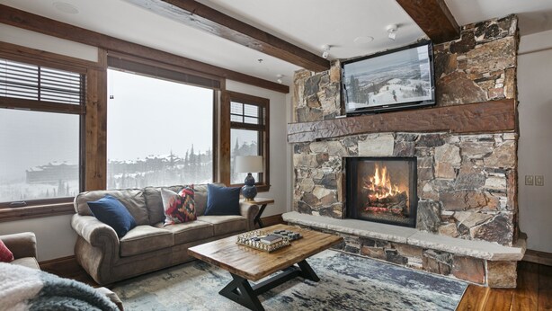 A cozy living room with a stone fireplace, a large TV mounted above it, and a view of a snowy landscape through large windows. - Grand Lodge - Three Bedroom Condo #401 - Ski-in/ski-out by Deer Valley Resort (Park City)