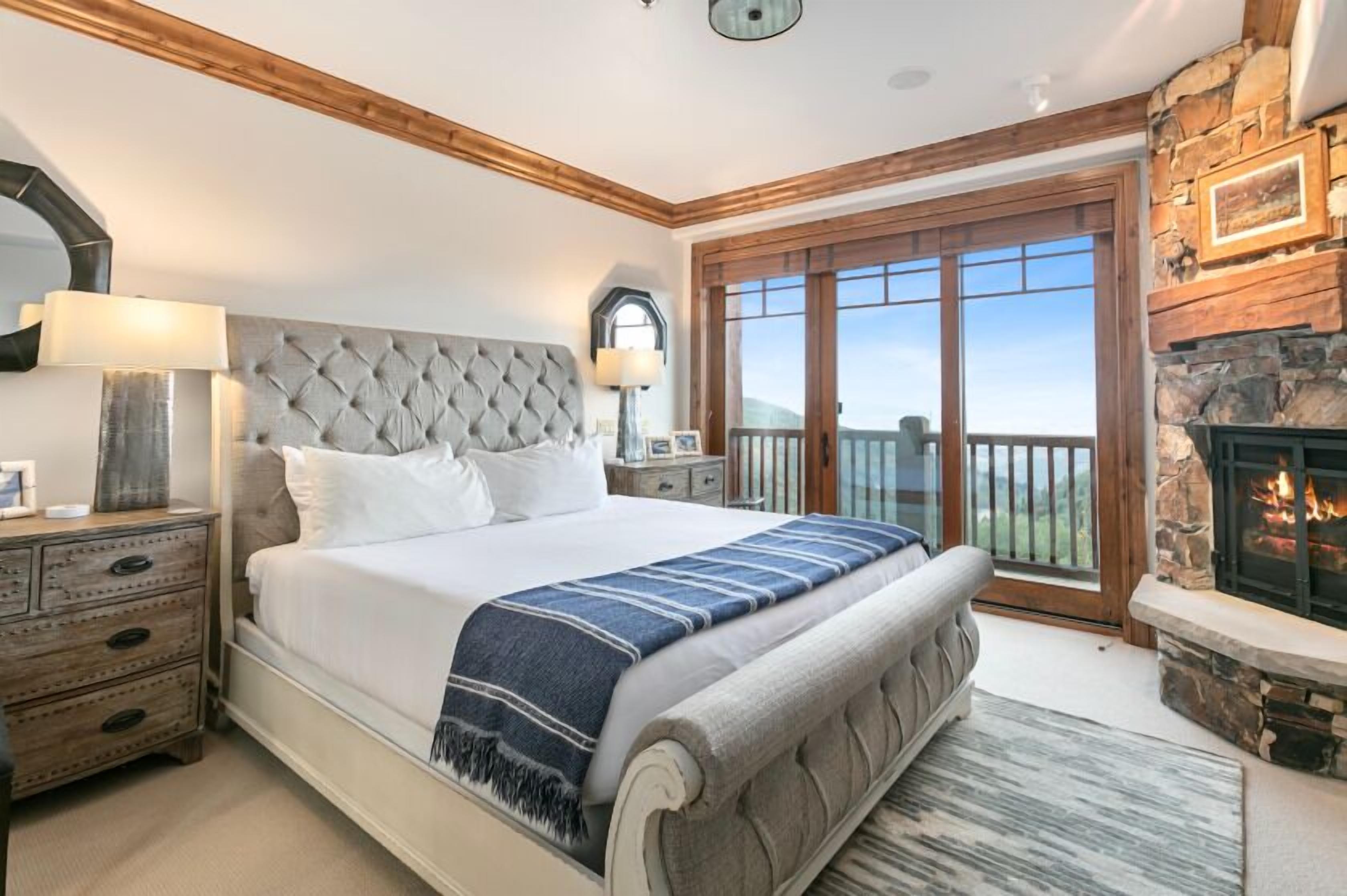A bedroom with a large bed, a stone fireplace, and a balcony with a view.