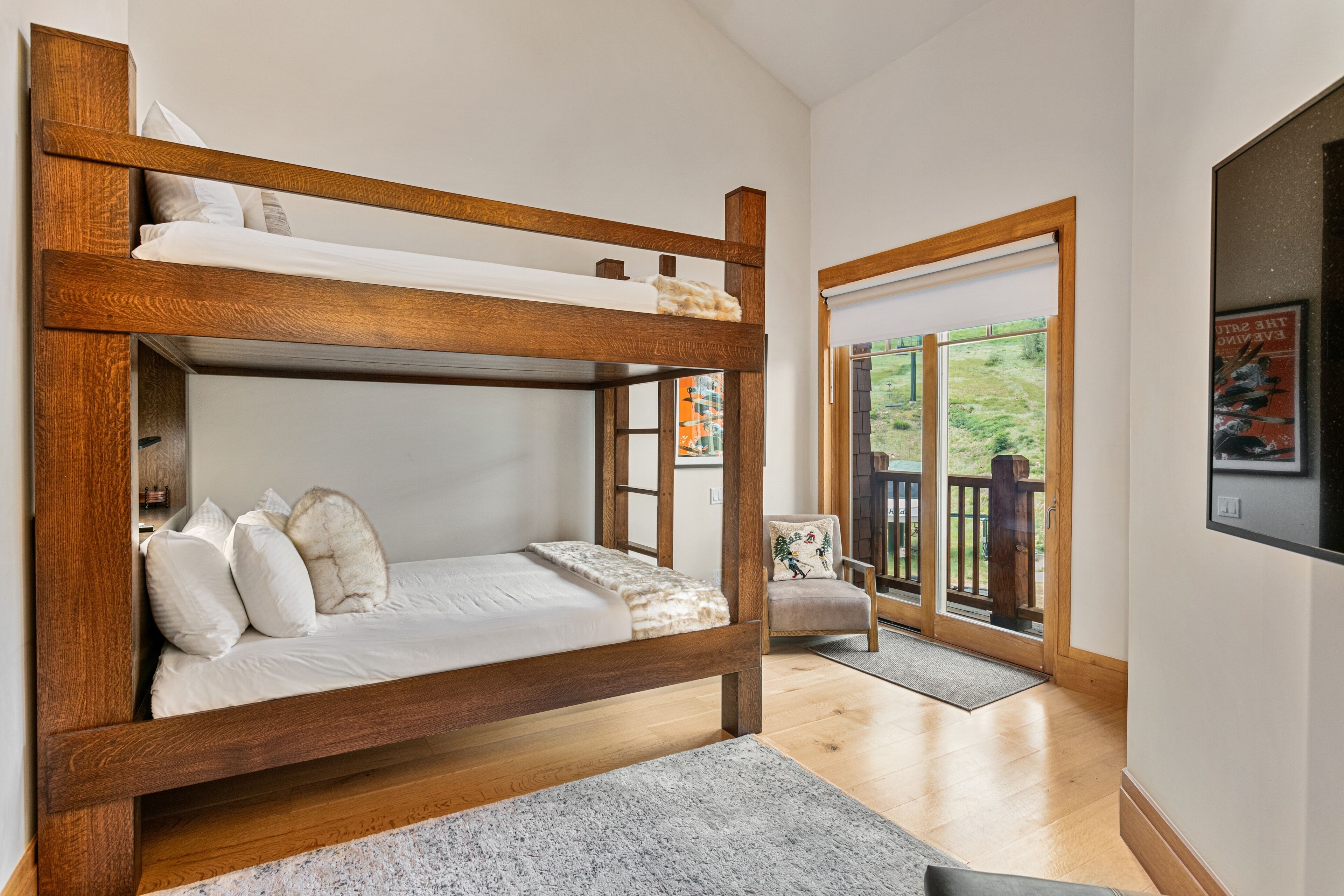 A bunk bed with a wooden frame, a white mattress, and several pillows. There is a chair with a cushion and a door leading to an outdoor area with a wooden deck.