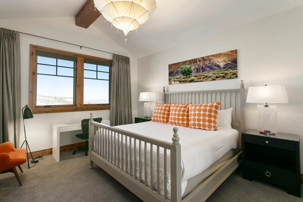 Una habitación de hotel con una cama grande, dos mesitas de noche con lámparas, un escritorio, una silla naranja y una ventana con cortinas.
