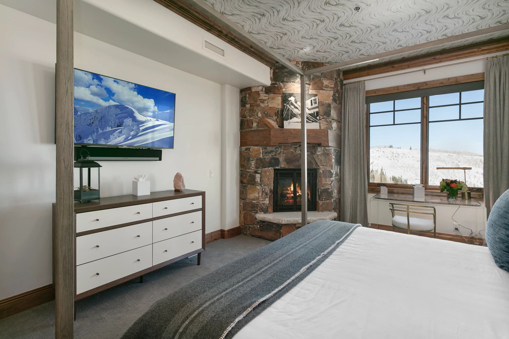 A bedroom with a stone fireplace, a large window with a view, a flat-screen TV, and a dresser.
