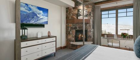 A bedroom with a stone fireplace, a large window with a view, a flat-screen TV, and a dresser.