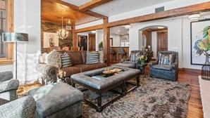 Un salon spacieux avec un grand canapé en cuir, une table basse et un lustre. La pièce est dotée de poutres en bois au plafond et d’un mur en pierre.