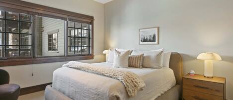 A bedroom with a large bed, a nightstand with a lamp, a chair, and a window with a view of buildings.