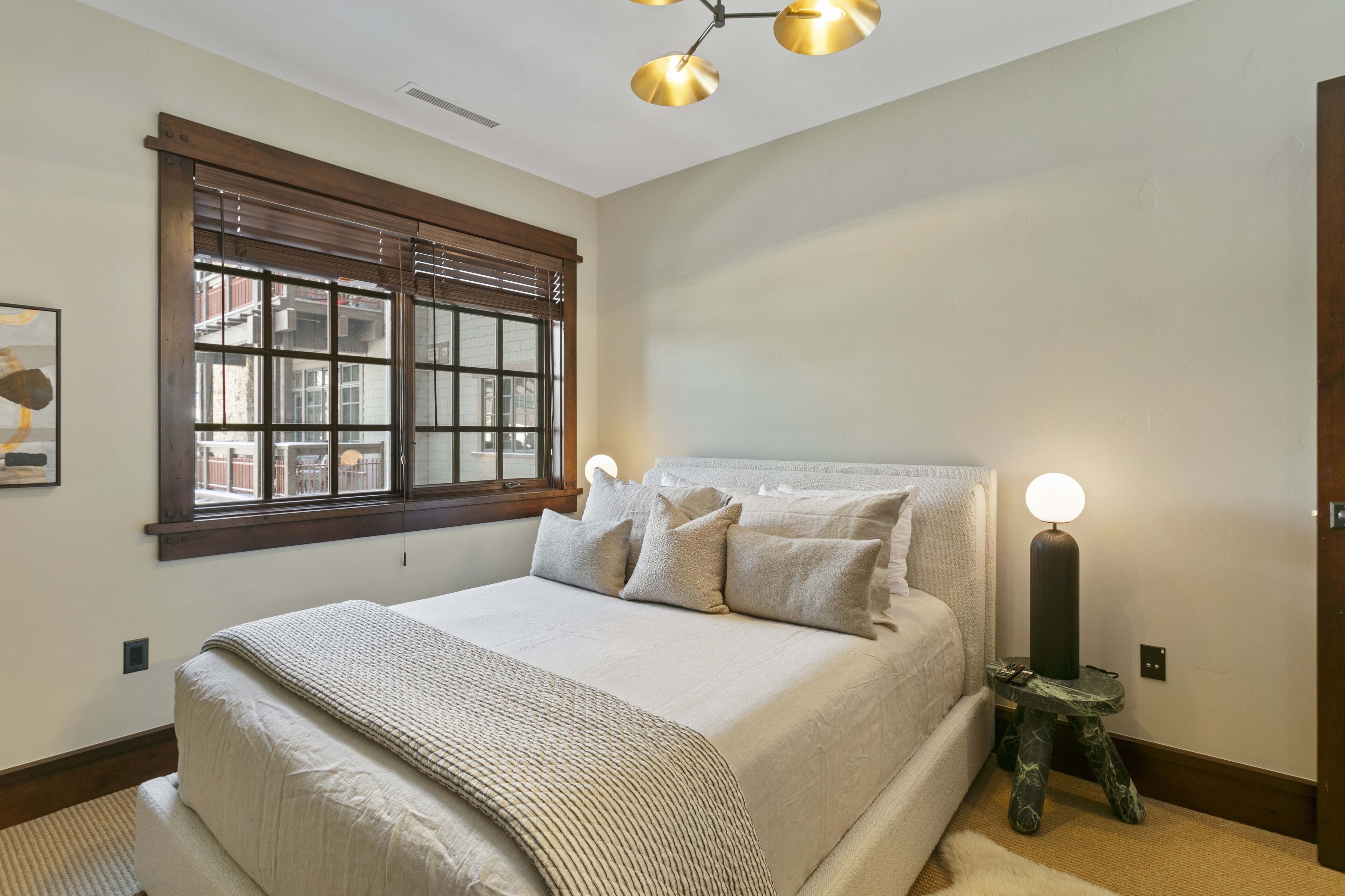 A bedroom with a large bed, a window with blinds, a bedside table with a lamp, and a ceiling light fixture.