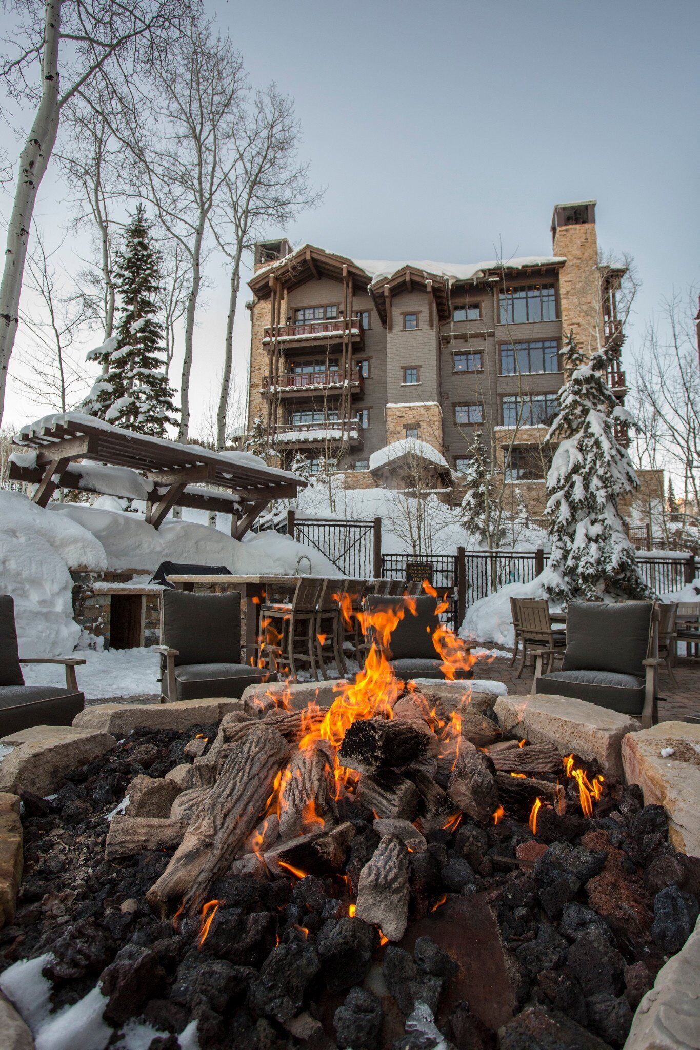 A fire pit with logs in a stone surround, surrounded by outdoor seating and snow-covered trees.