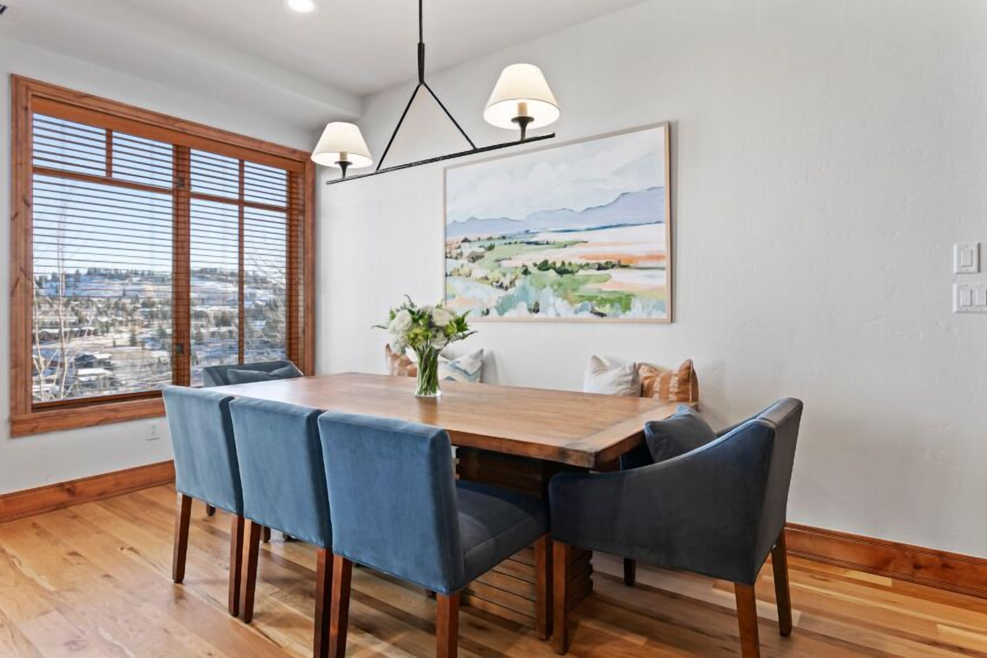 Un comedor con mesa de madera y sillas azules, un cuadro en la pared y una ventana con persianas.