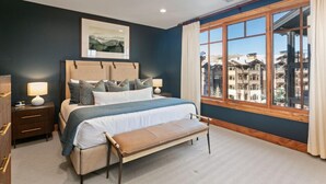 A bedroom with a large bed, a bench, a nightstand, a framed picture, and a view of buildings through a window. - 305 - Three Bedroom Condo Shooting Star by Deer Valley Resort (Park City)