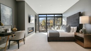 A modern bedroom with a large bed, a desk, and a chair. There is a fireplace and a large window with a view of trees and buildings. - 201 - Four Bedroom Argent 4 Condo by Deer Valley Resort (Park City)