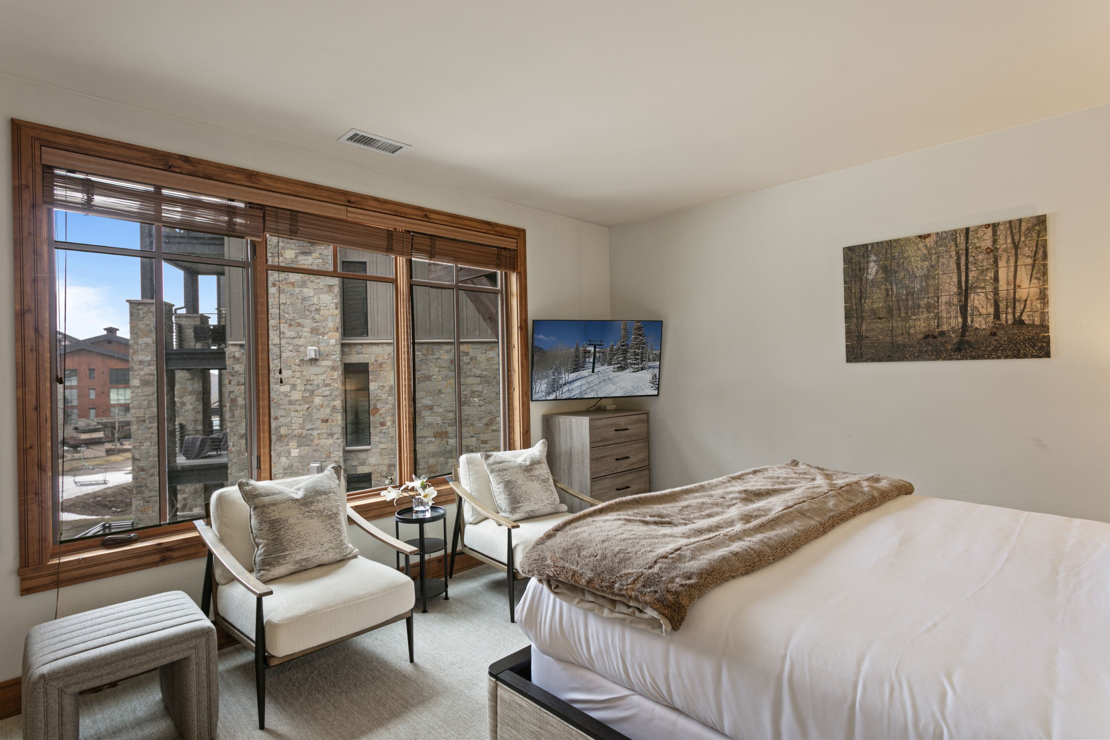 A bedroom with a large window, a bed, a chair, a small table, and a painting on the wall.