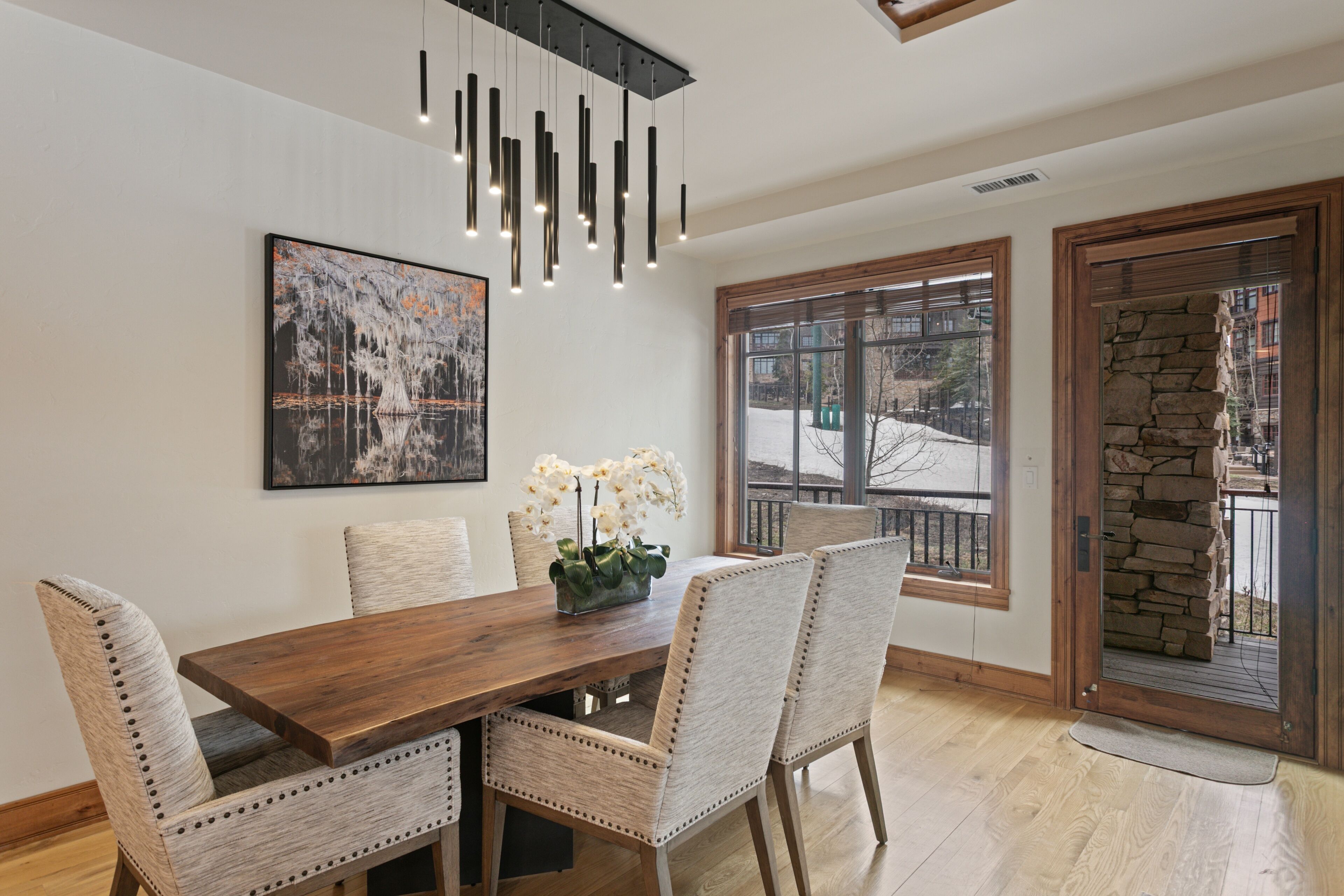Un comedor con mesa de madera y sillas acolchadas, lámpara colgante y un ventanal con vistas al exterior.