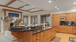 A modern kitchen with wooden cabinets, a large island, stone fireplace, and a scenic view through the windows. - 106 - Three Bedroom Condo Shooting Star by Deer Valley Resort (Park City)