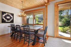 A dining area with a wooden table and chairs, a chandelier, and a large window with a view of greenery.