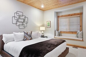 A bedroom with a wooden ceiling, a bed with white bedding and a dark throw, a bedside table with a lamp, a window with blinds, and a wall art piece.