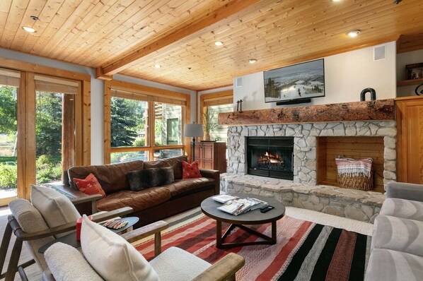 A cozy living room with a stone fireplace, wooden ceiling, and large windows overlooking a forest.