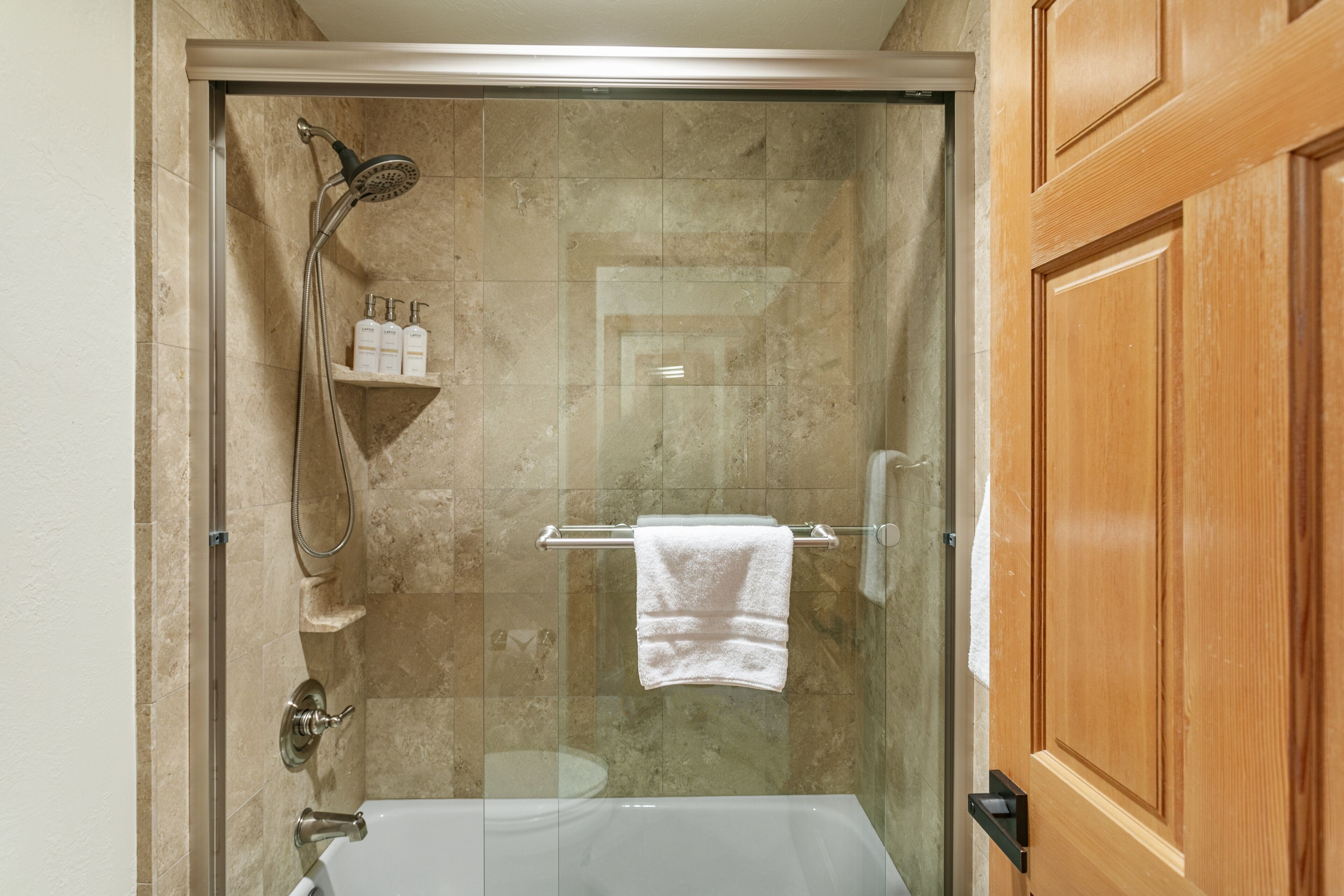 Una ducha con mampara de vidrio, cabezal de ducha manual y una bañera con mampara de vidrio, junto a una puerta de madera.