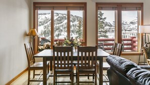 Un área de comedor con una mesa y sillas de madera, un jarrón de flores y ventanas amplias con vistas a una montaña nevada.