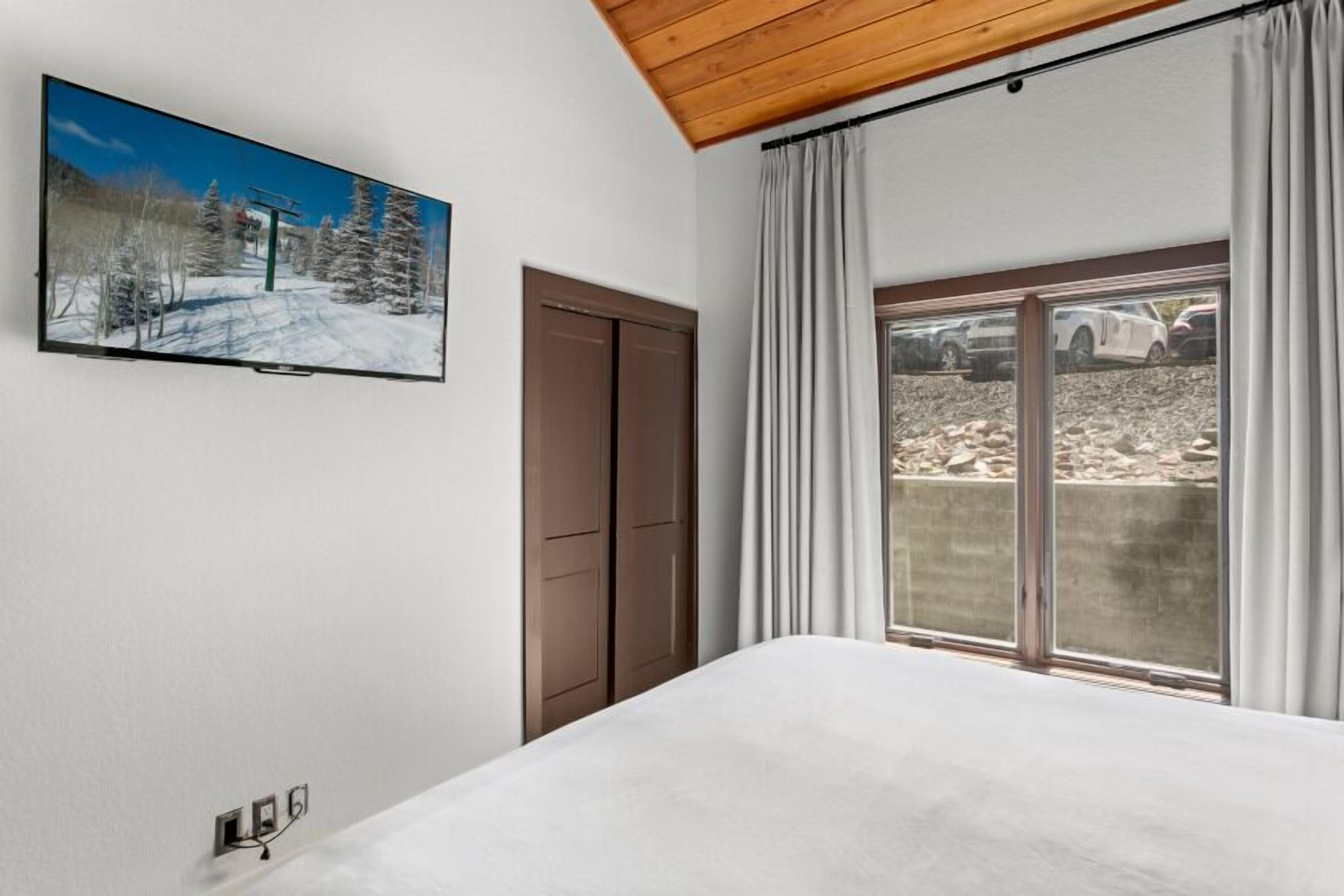 Une chambre avec un grand lit, une porte-fenêtre coulissante donnant sur un balcon, un téléviseur fixé au mur et un plafond en bois.
