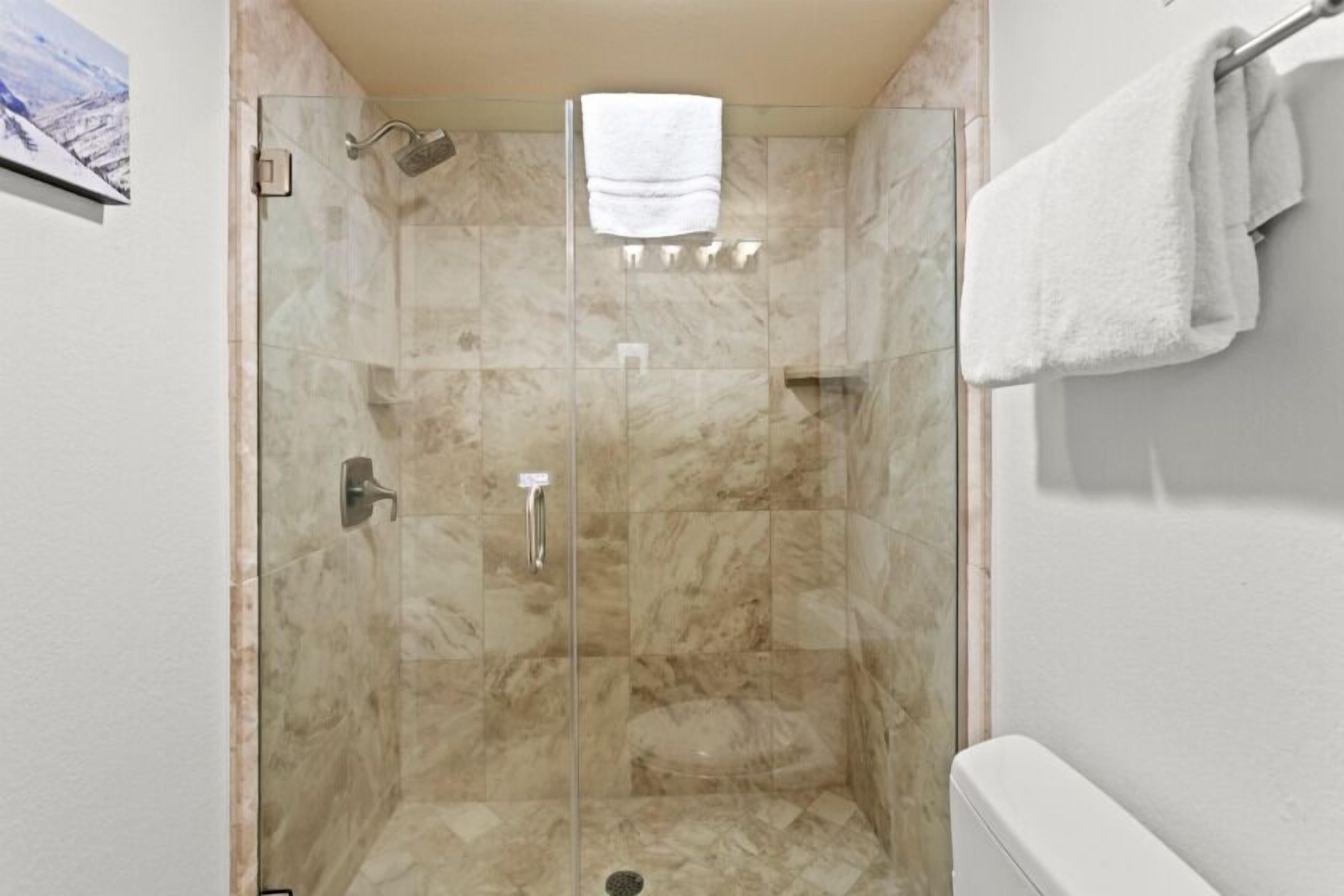 A glass-enclosed shower with a marble floor, a white towel hanging on the wall, and a white bathtub.