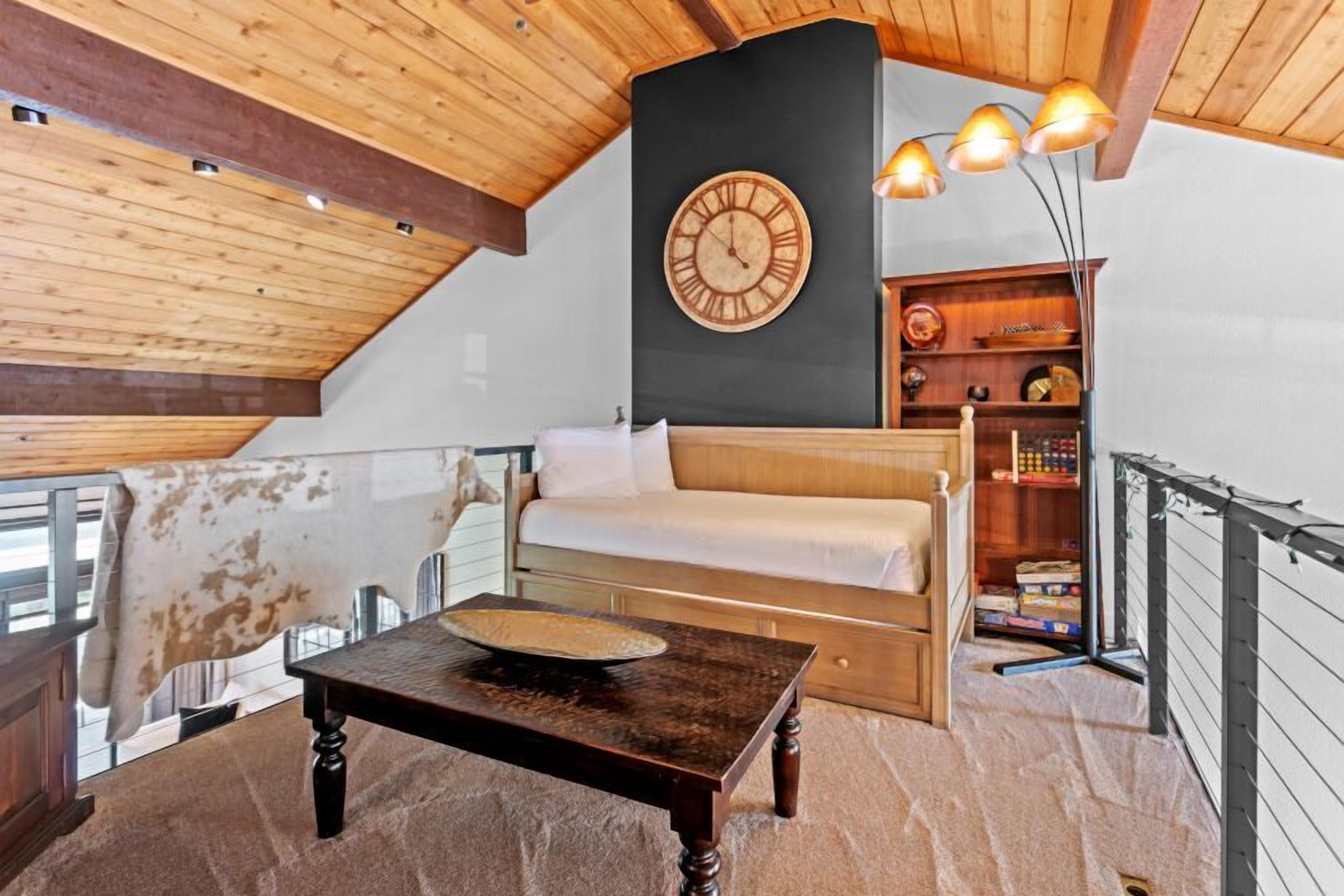 A cozy loft bedroom with a wooden ceiling, a large wall clock, a sofa, a small table, and a bookshelf.