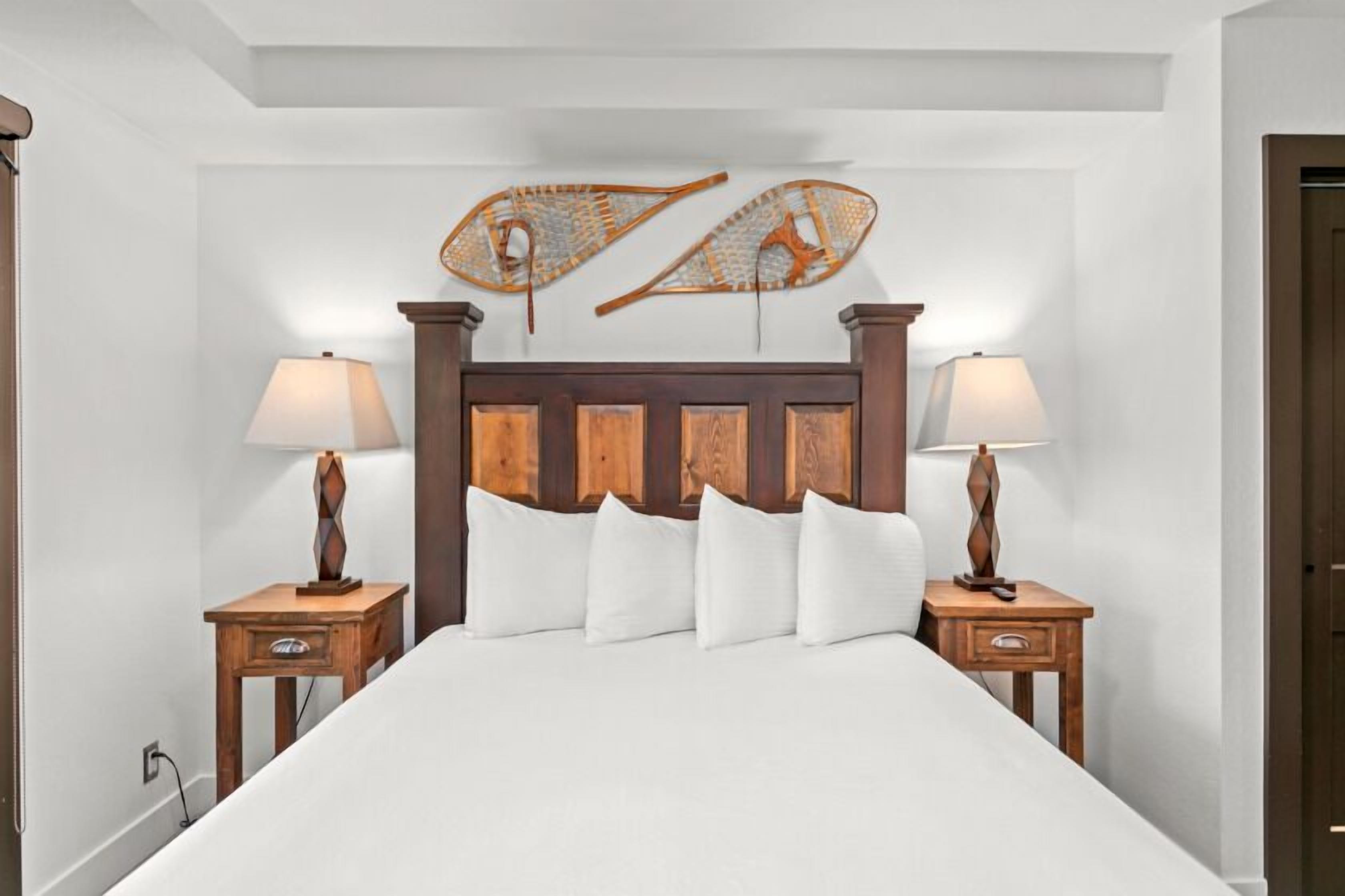 Un dormitorio ordenado con cabecera de madera, ropa de cama blanca, dos mesitas de noche con lámparas y decoración en la pared.