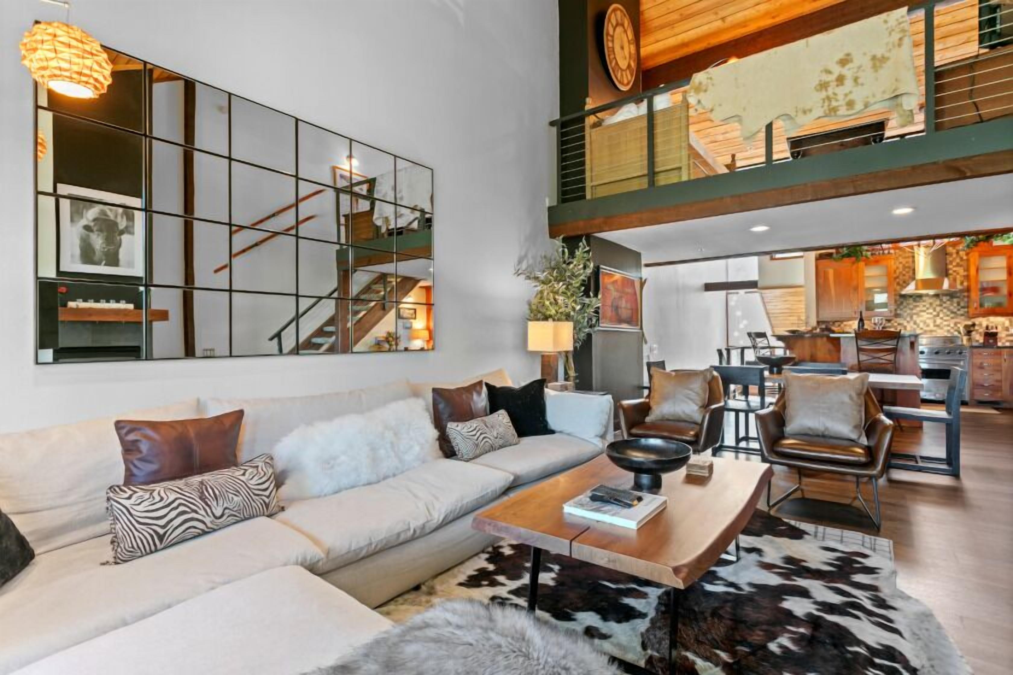 A spacious living area with a sofa, coffee table, and a large mirror. A staircase leads to an upper level with a kitchen visible.