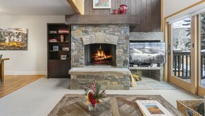 A cozy living room with a stone fireplace, a bookshelf, and a large window with a view of trees. - 1490 - Three Bedroom Condo with Spa Aspenwood by Deer Valley Resort (Park City)
