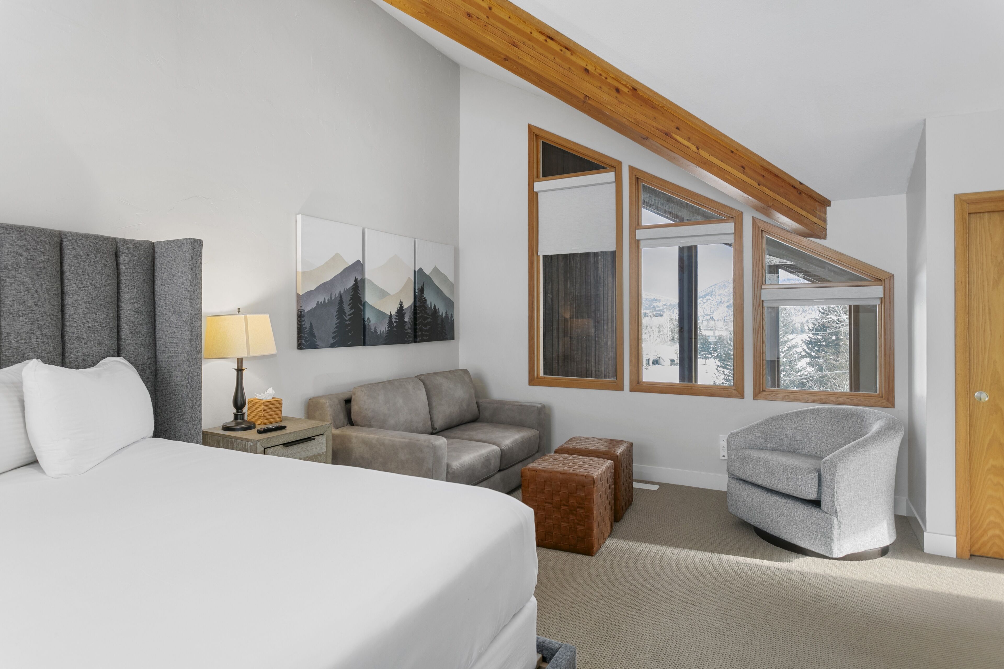 A modern hotel room with a large bed, a gray sofa, a gray armchair, and a wooden ceiling beam.