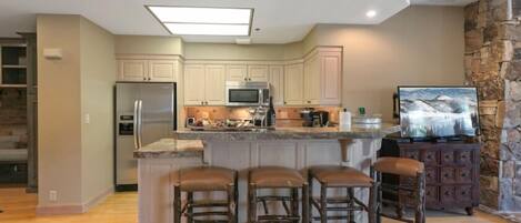 A modern kitchen with a central island, bar stools, a stone accent wall, and a built-in TV.