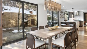 A modern dining area with a large table and chairs, a chandelier, and a view of a snowy landscape through large windows. - 656 Coalition View Court - 4 Bedroom Private Home With Spa by Deer Valley Resort (Park City)
