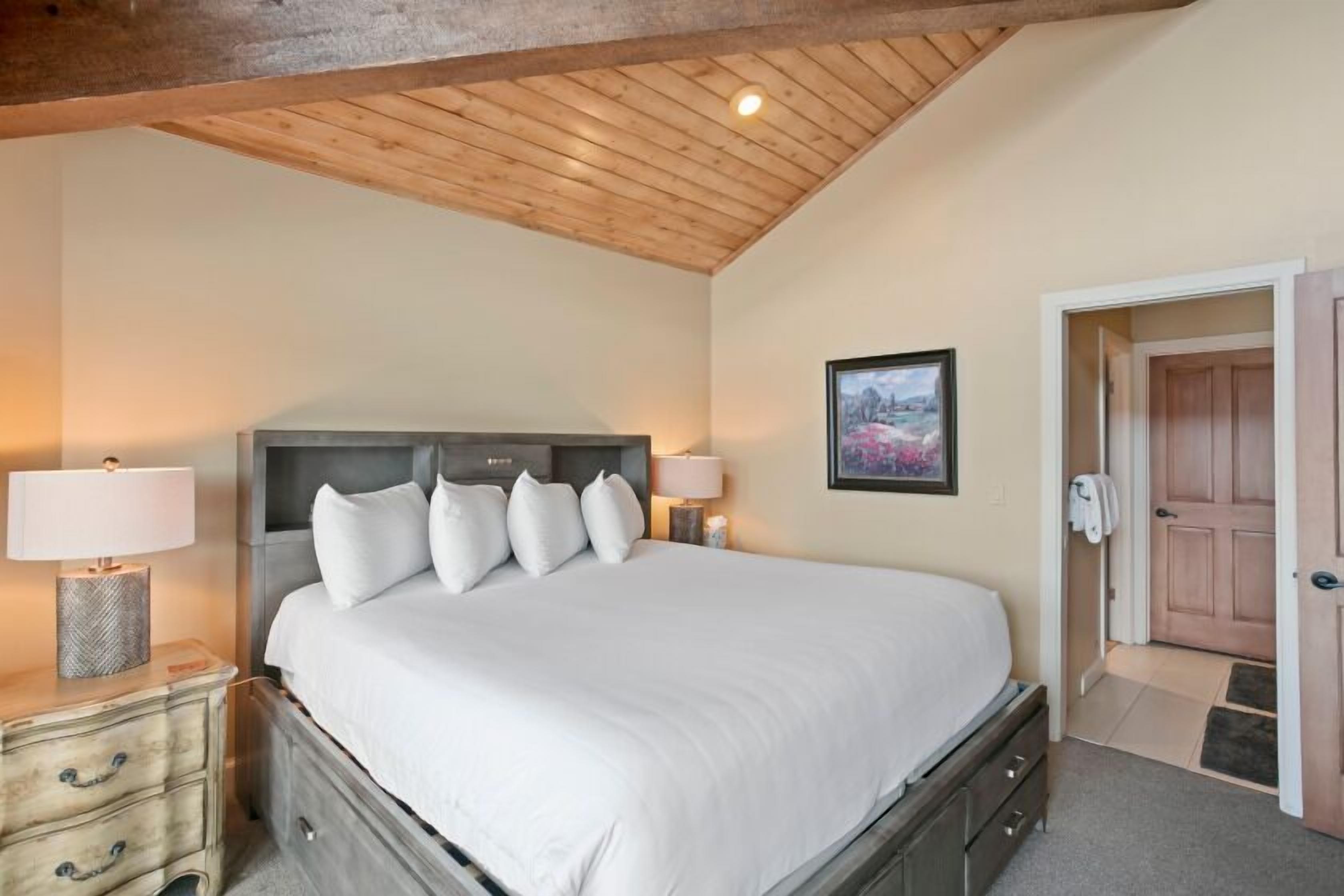 A bedroom with a large bed, wooden headboard, two bedside tables with lamps, a framed picture on the wall, and a wooden door.