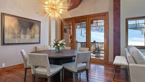 A dining area with a round table, six chairs, a chandelier, a large framed picture, and a view of a snowy landscape through glass doors. - 504 - Five Bedroom Condo Penthouse Grand Lodge by Deer Valley Resort (Park City)