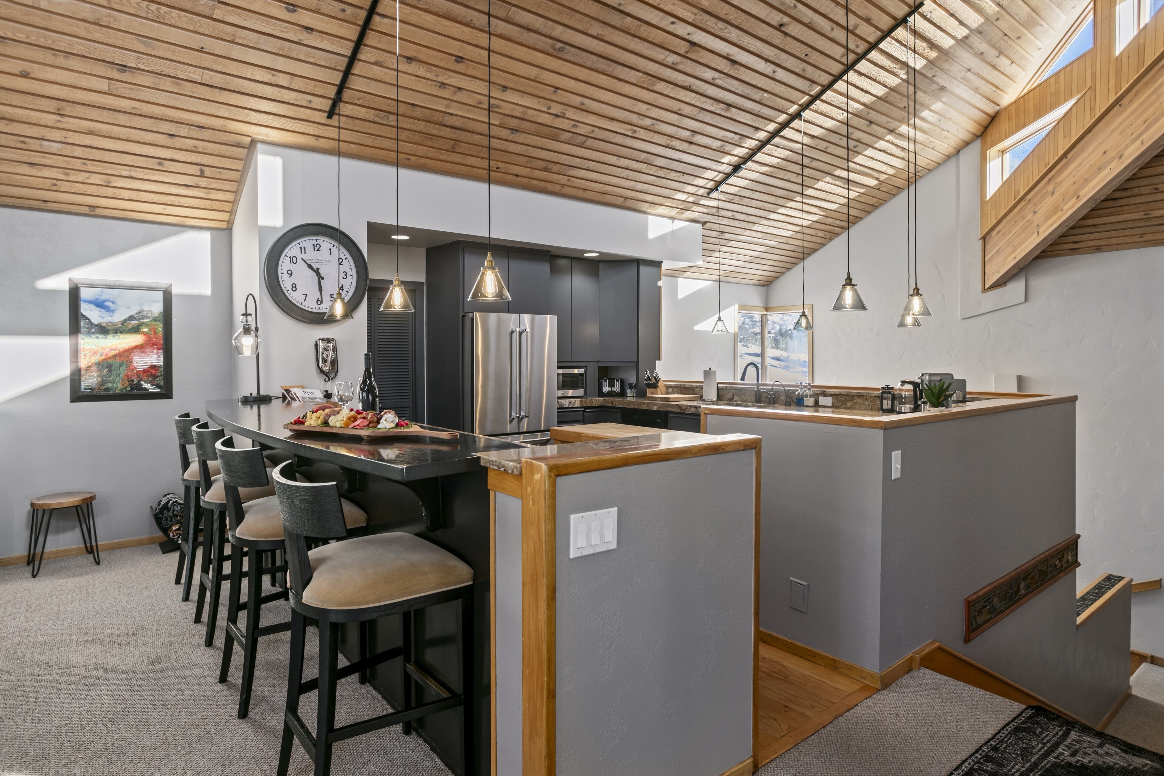 Una cocina moderna con una isla central, taburetes de bar, iluminación colgante y un reloj en la pared.