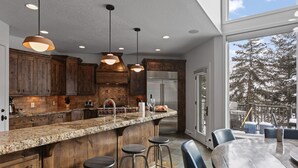 A modern kitchen with wooden cabinets, a granite countertop, and stainless steel appliances. There are bar stools and a dining table with chairs. - 1688 - Five Bedroom Condo with Hot Tub Pinnacle by Deer Valley Resort (Park City)
