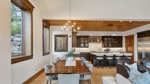 Una cocina moderna con vigas de madera, un comedor con una larga mesa y sillas, y una sala de estar con sofá y cojines.