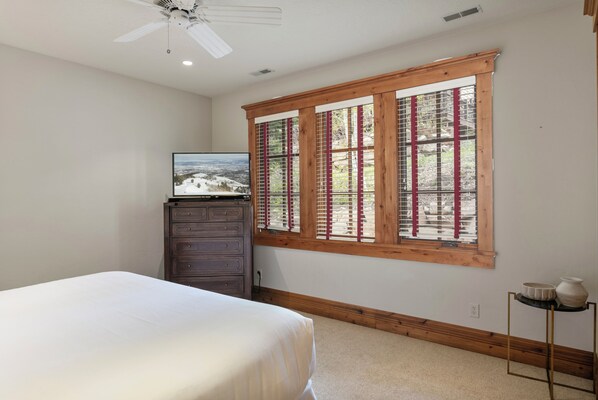 A bedroom with a bed, a dresser, a television, a window with wooden frames, and a small table with decorative items. - 6565 Lookout Drive - Four Bedroom Condo with Spa by Deer Valley Resort (Park City)
