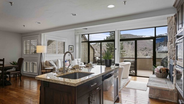A modern kitchen with a granite countertop, dark cabinetry, and a large island with a sink. There is a dining area with a table and chairs, and a view of a mountainous landscape through large windows.