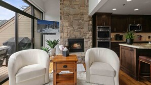 A modern living room with a stone fireplace, two white armchairs, a wooden coffee table, and a kitchen with dark wood cabinets and stainless steel appliances.