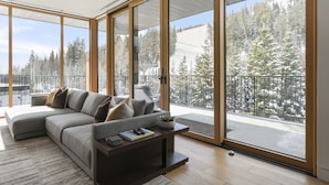 A modern living room with a sectional sofa, a coffee table, and large sliding glass doors leading to a balcony with a snowy mountain view. - 1504 Founders Place - Three Bedroom Condo Deluxe by Deer Valley Resort (Park City)