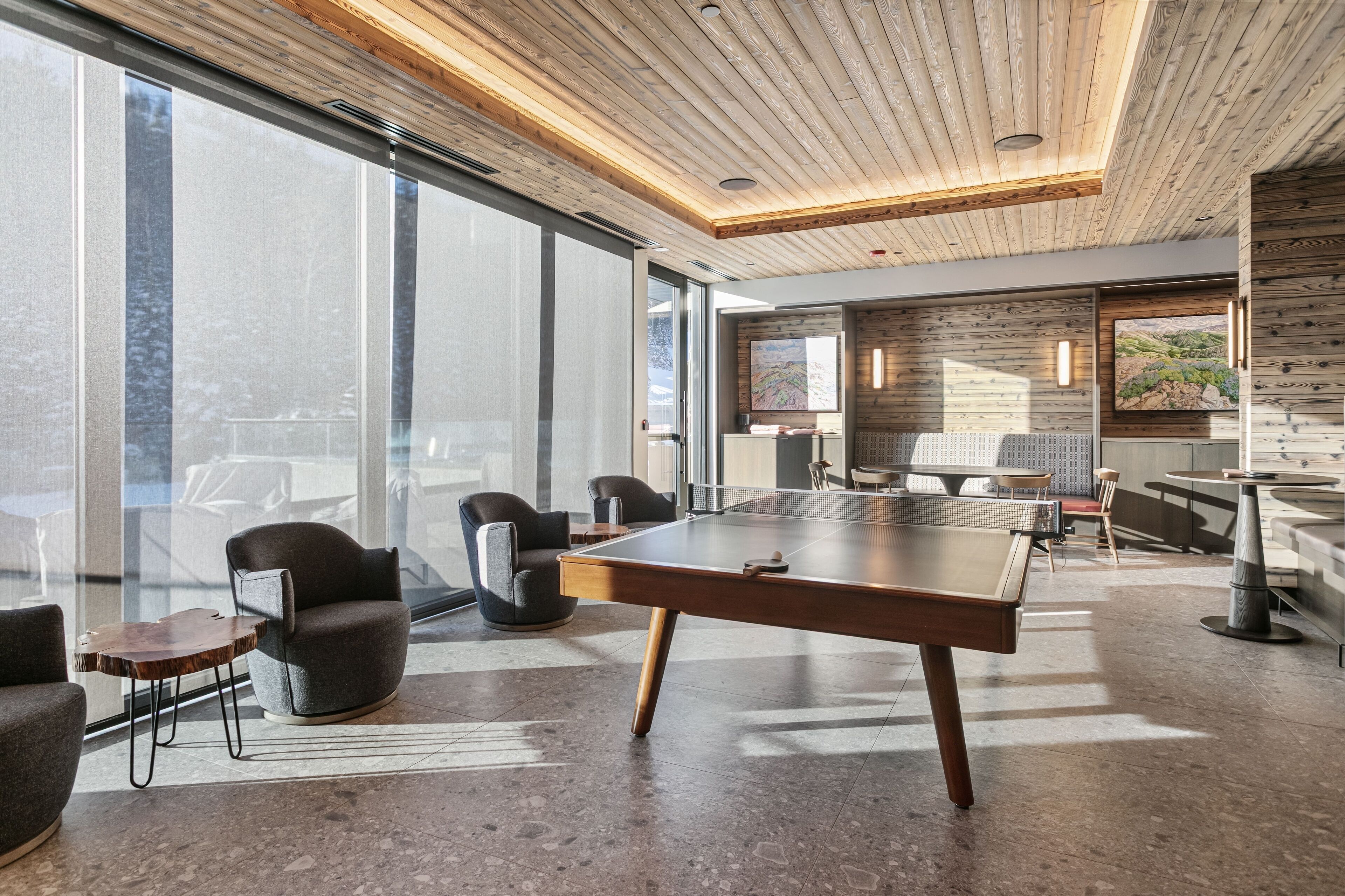 Un ambiente interno moderno con un tavolo da ping pong, comodi posti a sedere, ampie finestre e un soffitto in legno.