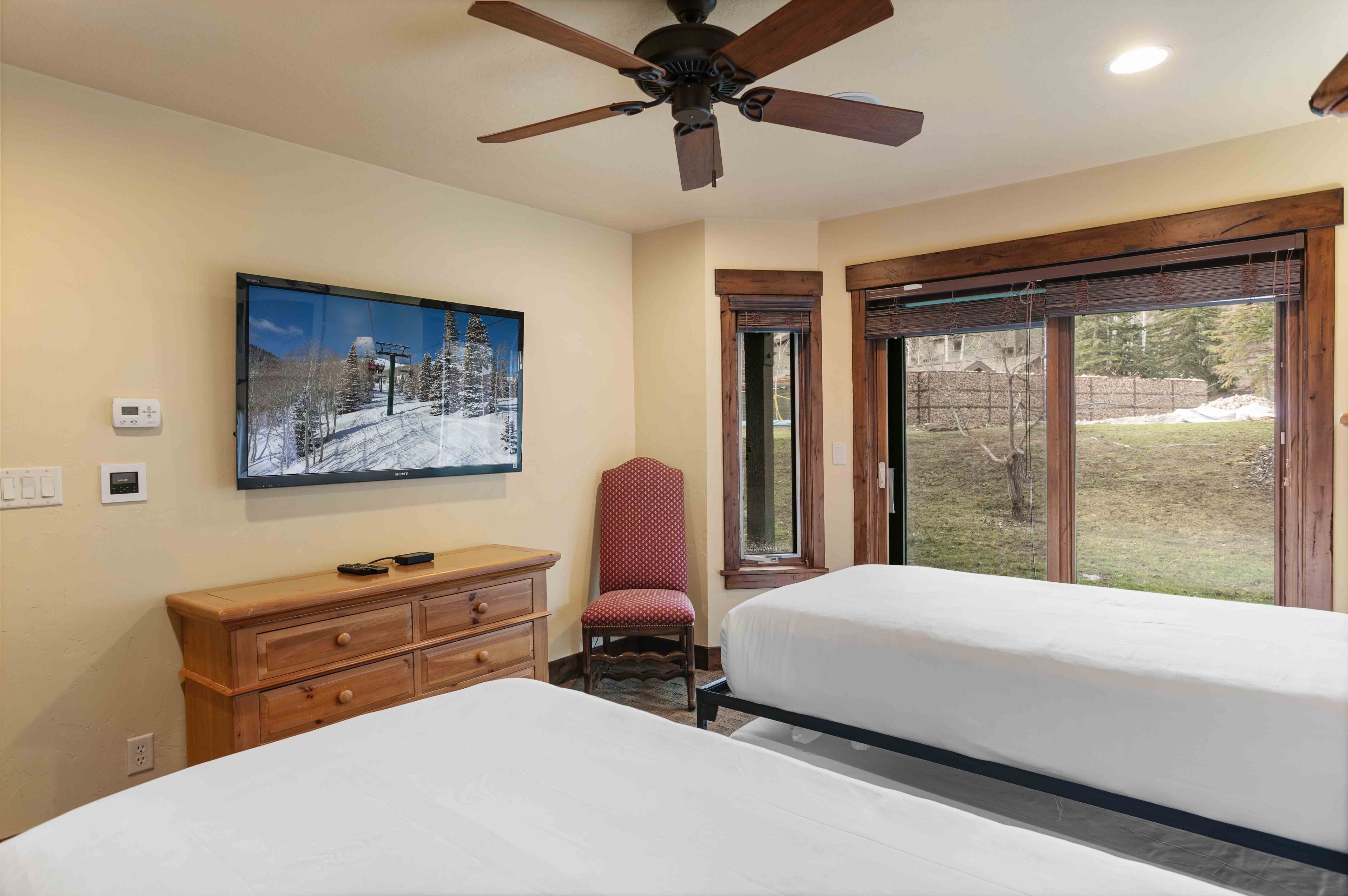 Une chambre d’hôtel avec deux lits, une commode en bois, un fauteuil à pois rouges, un ventilateur de plafond et une grande fenêtre donnant sur un paysage enneigé.