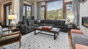 A modern living room with a large sectional sofa, a coffee table, and a fireplace. There are large windows offering a view of the outdoors.