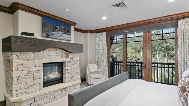 A bedroom with a stone fireplace, a large bed, a chair, and a balcony with a view of trees and a cabin.