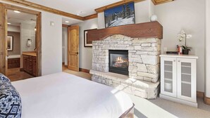 A bedroom with a stone fireplace, wooden ceiling beams, a bed, and a bathroom with wooden cabinets. - 302 - Four Bedroom Condo Silver Strike by Deer Valley Resort (Park City)